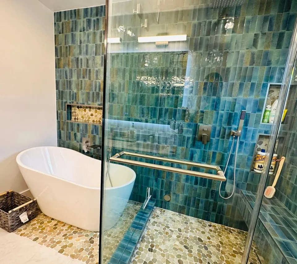 Remodeled primary bathroom with a white freestanding soaking tub, pebble-tile floor and a glass-enclosed shower in deep-teal vertical tile