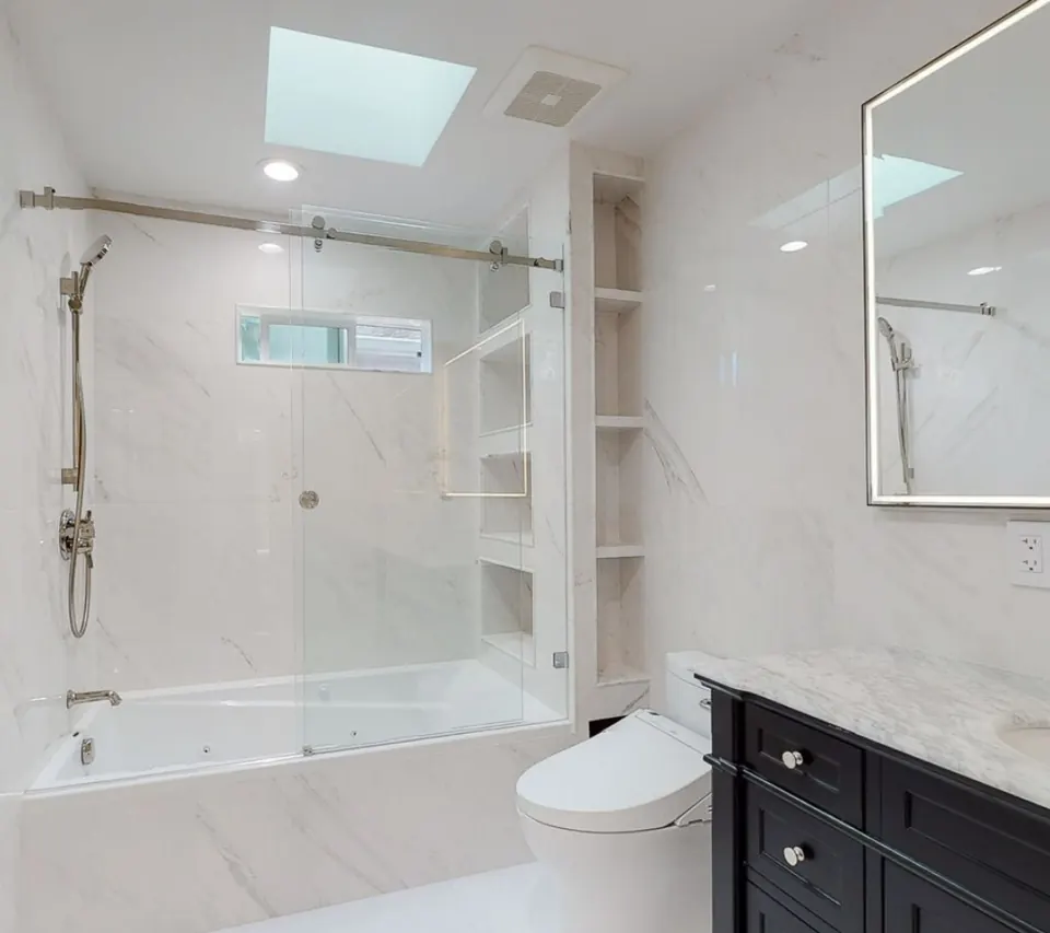 Remodeled bathroom with a marble-look tub and shower behind a sliding glass door, built-in shelving and a dark vanity