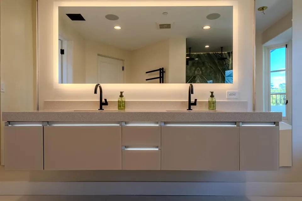 Taupe floating double vanity with matte-black faucets and a full-width backlit mirror reflecting a marble shower