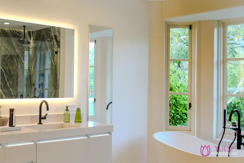 Primary bathroom with a backlit mirror, floating double vanity, matte-black faucets and a freestanding tub by the window