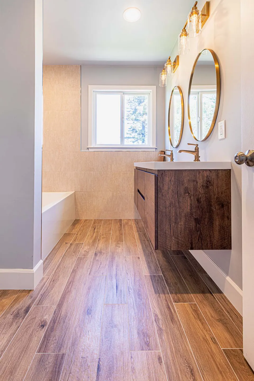 Primary bathroom with a walnut floating vanity, oval mirrors, wood-look plank tile and a soaking tub under the window