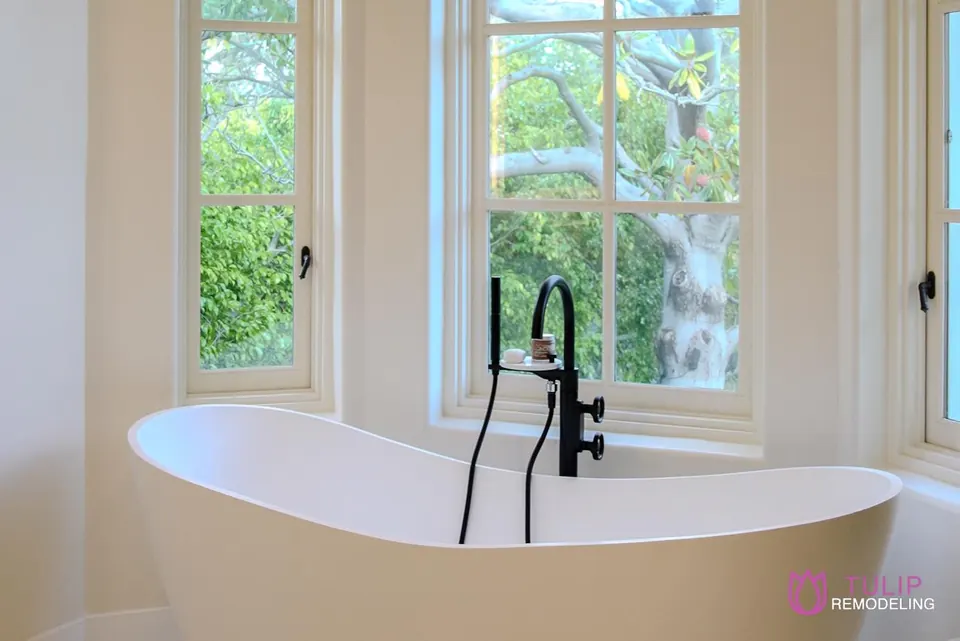 Freestanding soaking tub with a matte-black floor-mounted filler set beneath divided-light windows overlooking a garden