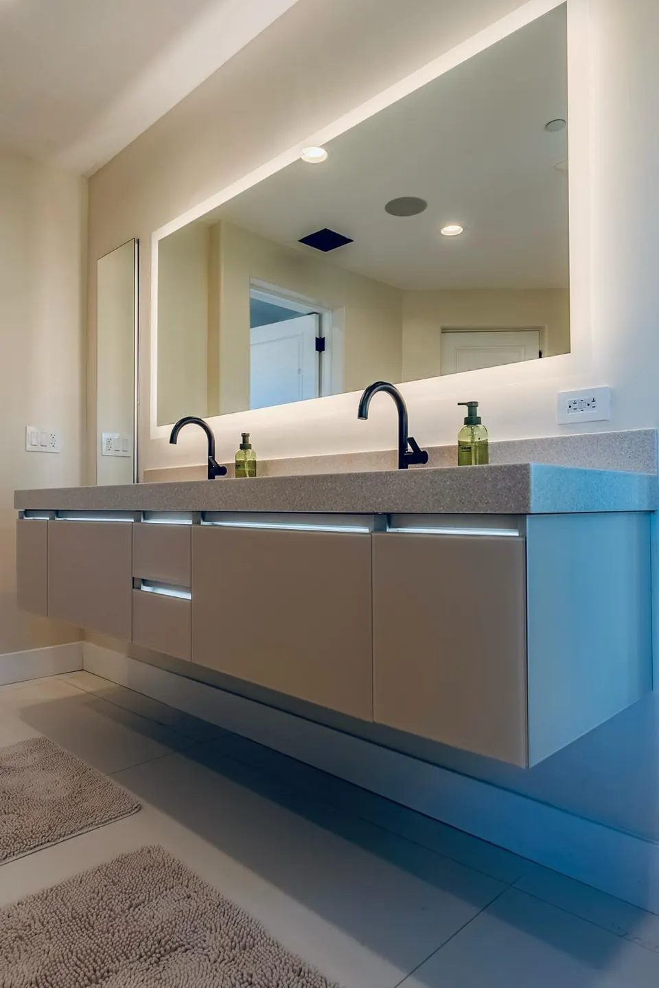 Floating vanity with toe-kick lighting, a quartz top, black faucets and a large backlit mirror