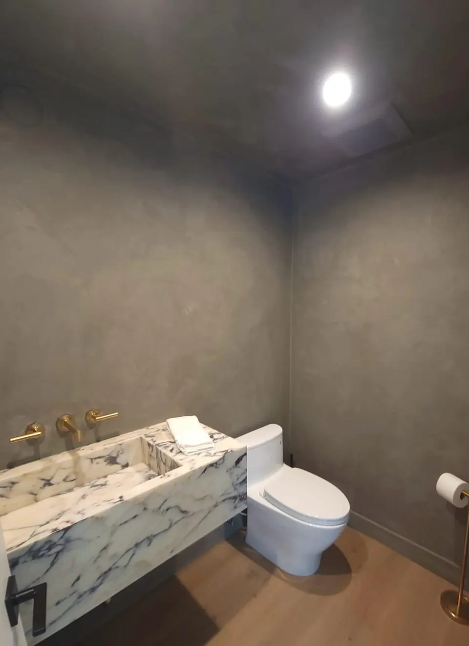 Powder room with a veined marble floating vanity, brass wall faucet, wall-hung toilet and gray plaster walls
