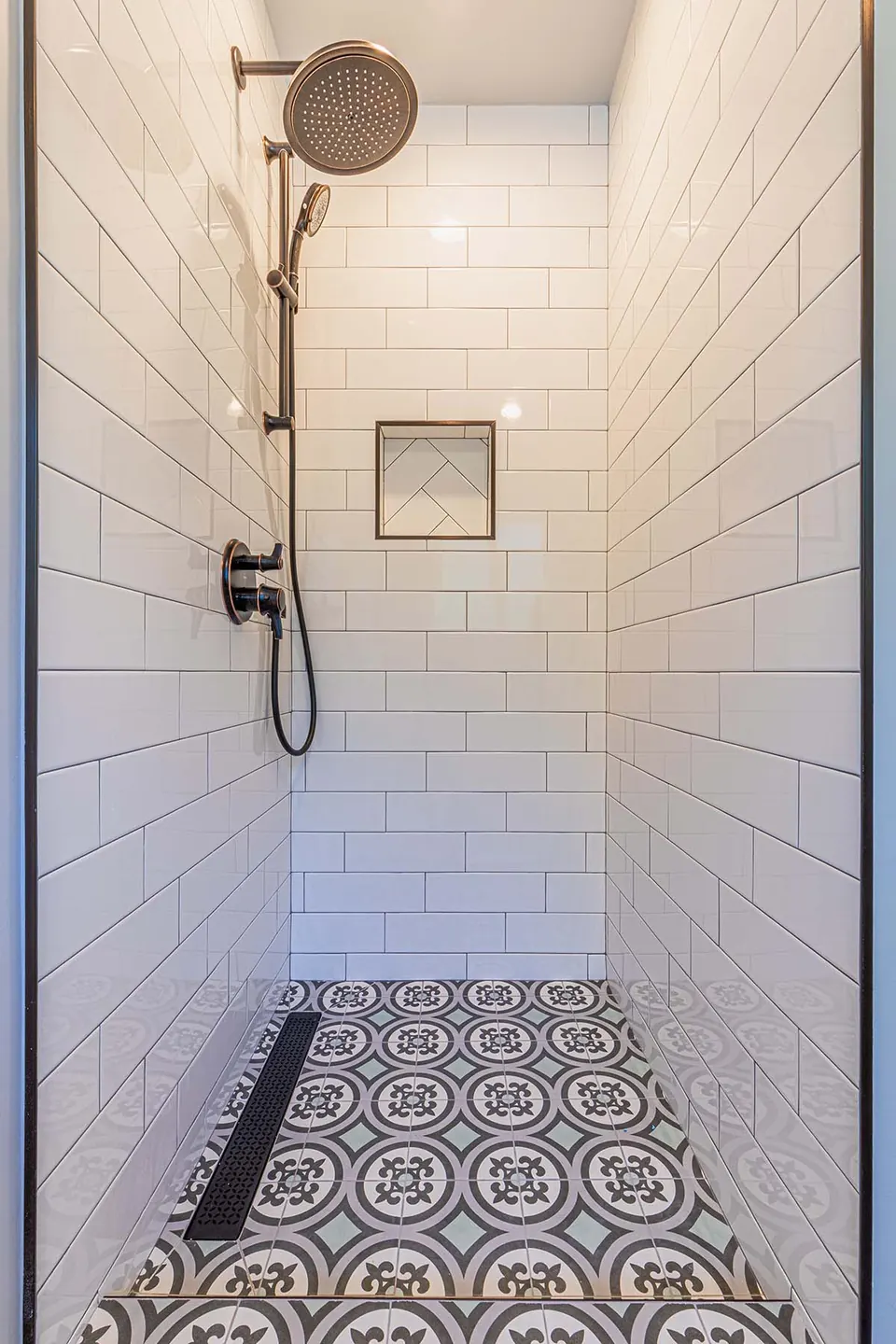 Walk-in shower in white subway tile with a recessed niche, matte-black rain head and a patterned floor with linear drain